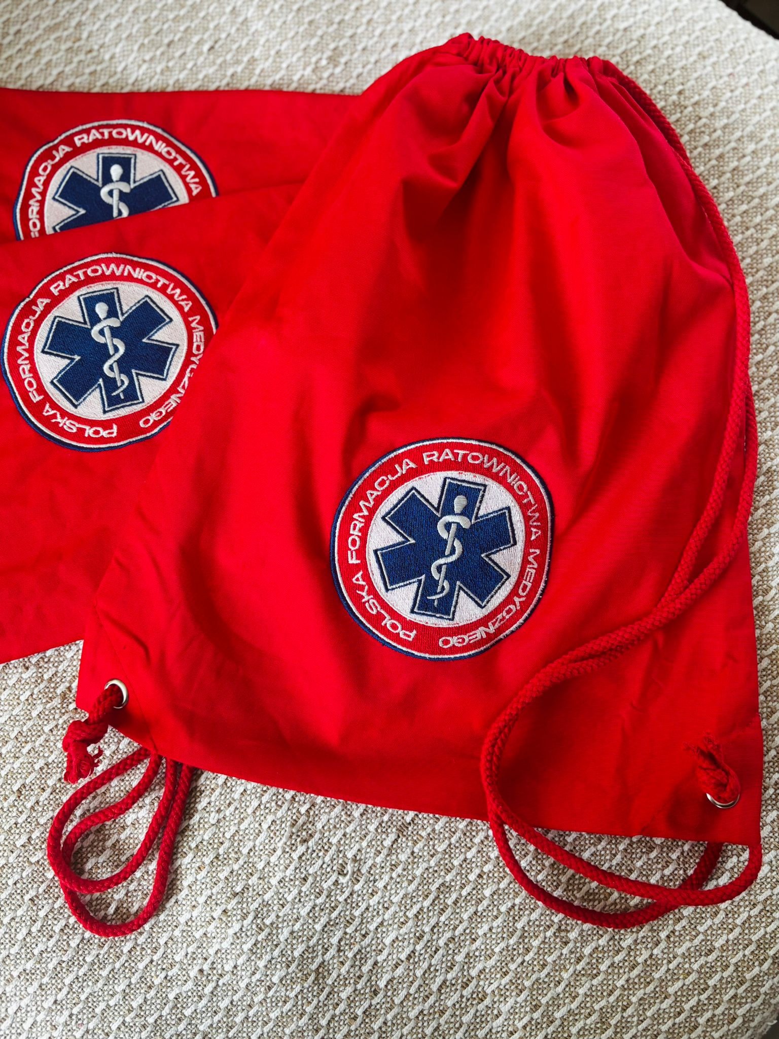 Czerwone worki z logo 'Formacja Ratownictwa Medycznego' i symbolem medycyny, leżące na teksturowanym tle. Widoczne detale sznurków i haftu.