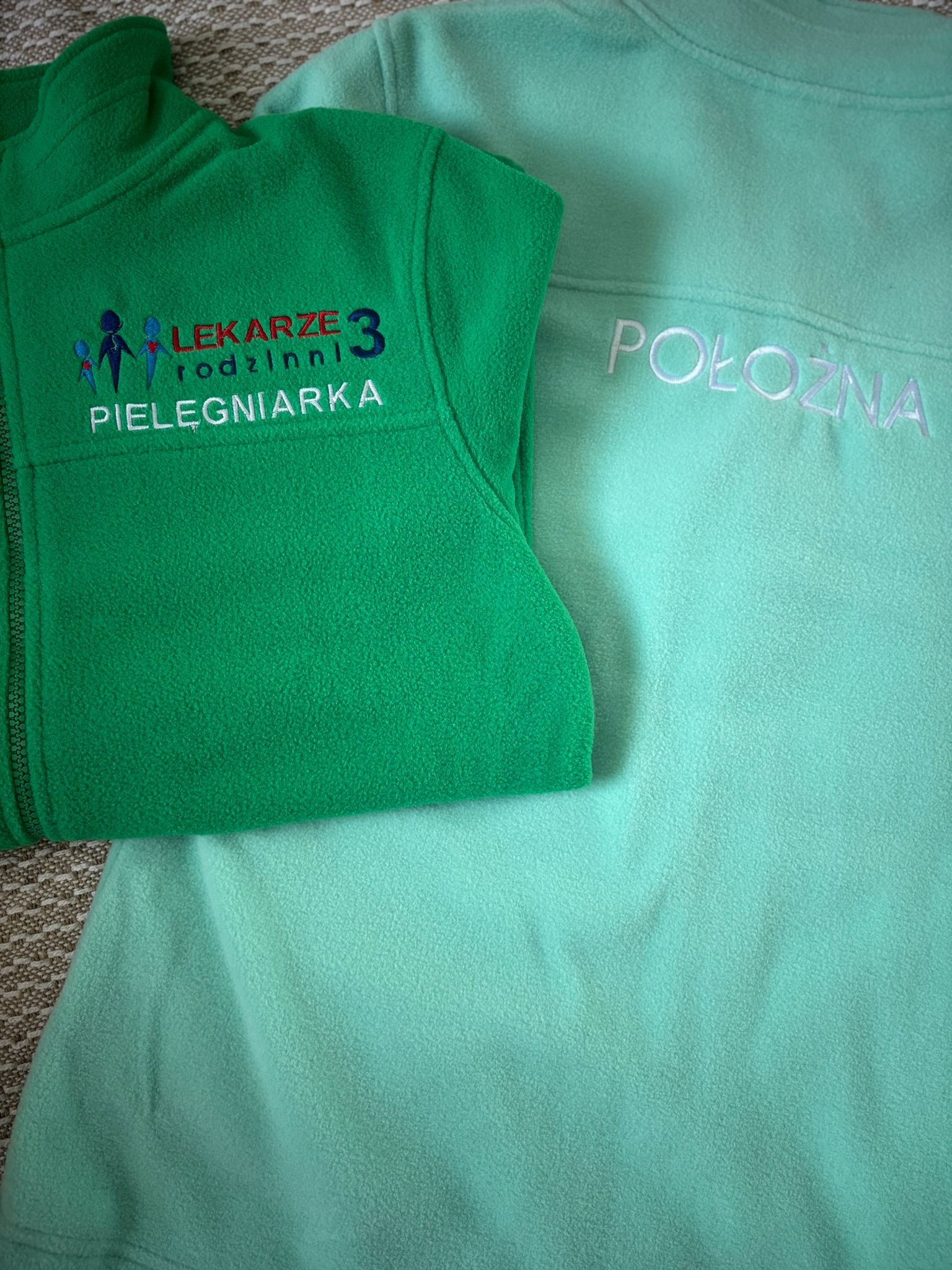 Zielona bluza polarowa z haftowanym napisem 'LEKARZE rodzinni PIELĘGNIARKA' i turkusowa bluza z napisem 'POŁOŻNA'.