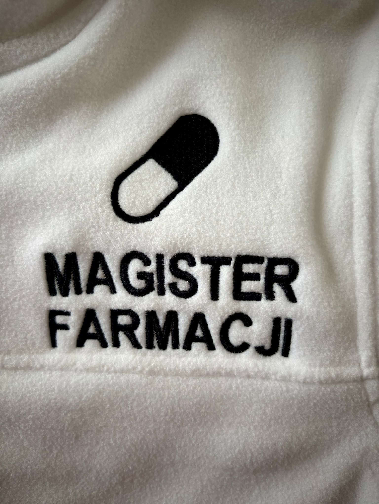 Biała bluza polarowa z czarnym, precyzyjnym napisem 'MAGISTER FARMACJI' i symbolem kapsułki, wykonanym techniką haftu.