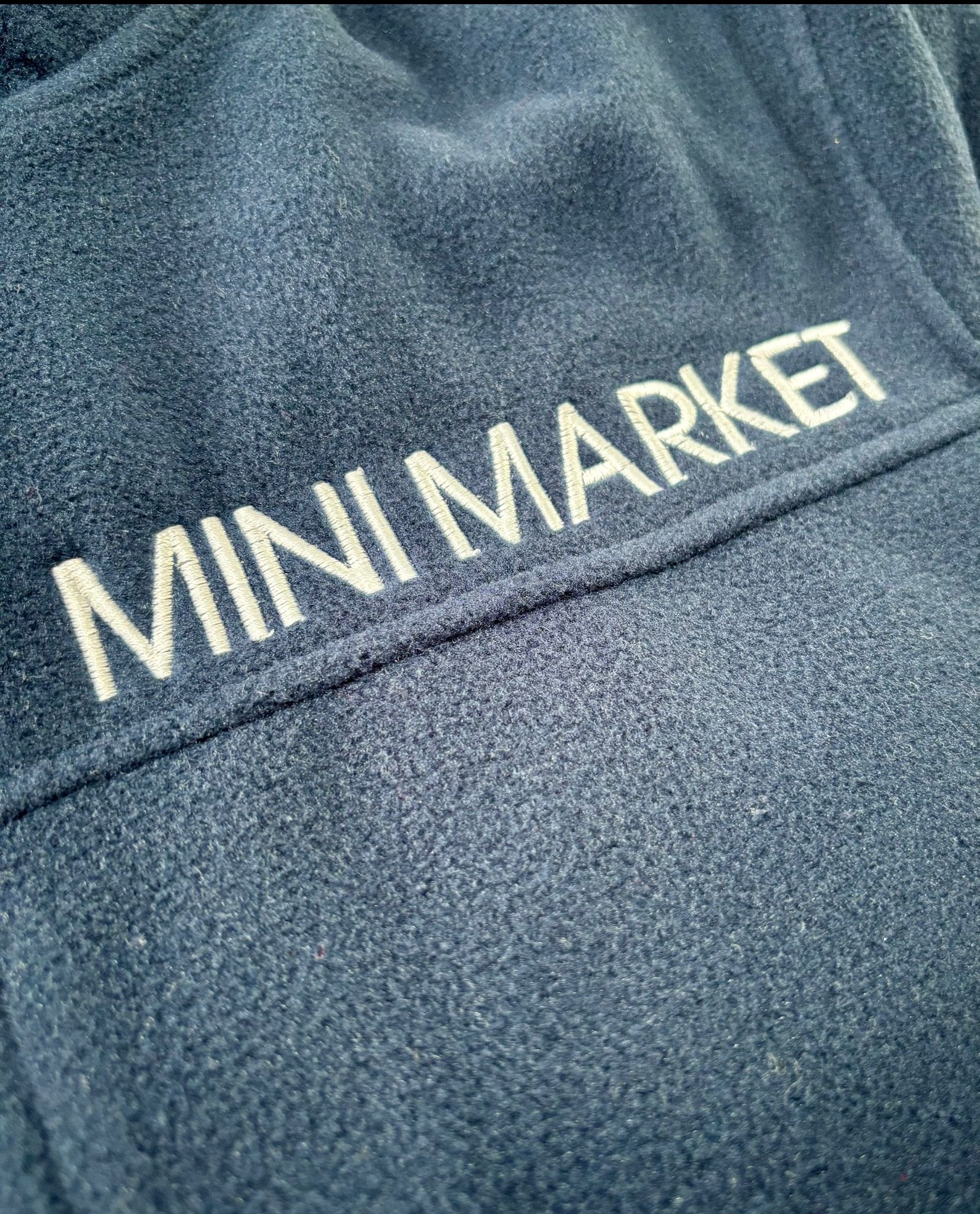 Granatowa tkanina z wyhaftowanym napisem 'MINI MARKET' białą nicią. Widoczna faktura materiału i precyzyjne wykonanie haftu.