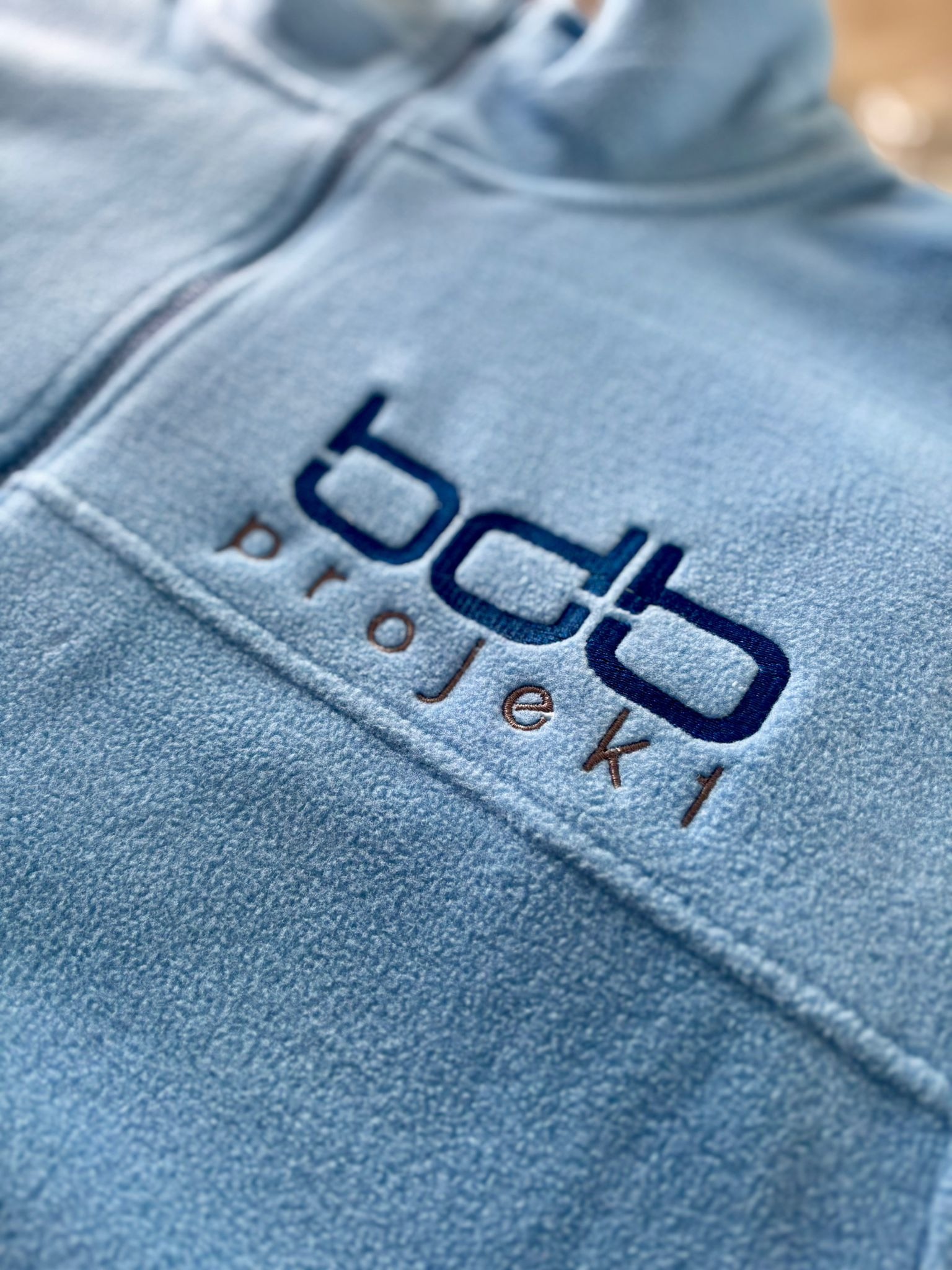 Jasnoniebieska bluza polarowa z haftowanym logo 'bcb projekty' w Żorach. Detal haftu, widoczna faktura materiału. Zamek błyskawiczny częściowo widoczny.