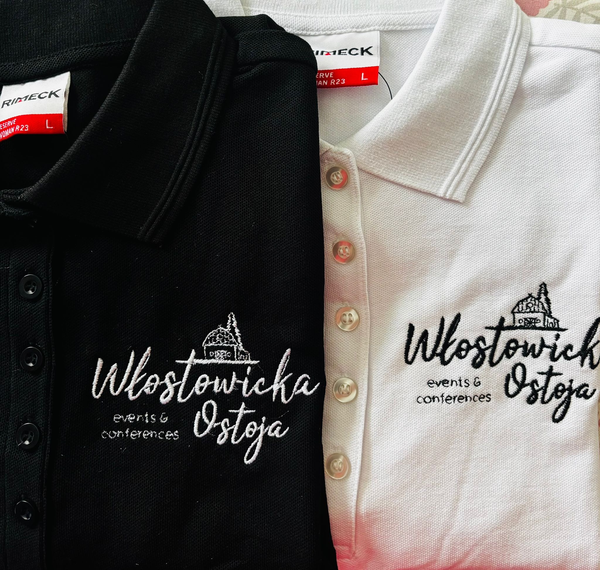 Dwie koszulki polo: czarna i biała, z wyhaftowanym logo firmy 'Włostowicka Ostoja events & conferences'. Widoczne guziki i metki.