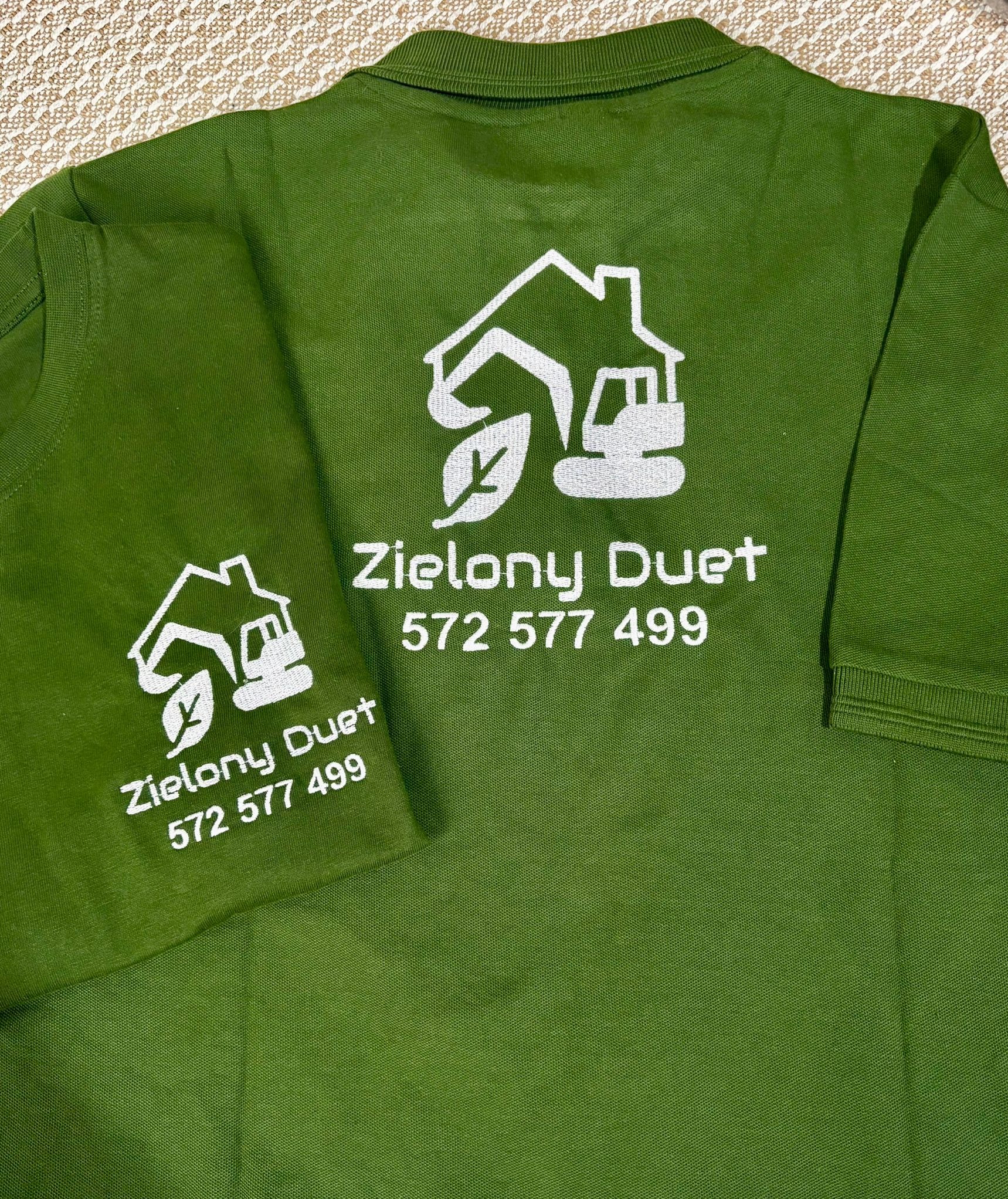 Zielona koszulka polo z haftowanym logo firmy 'Zielony Duet' i numerem telefonu, leżąca na teksturowanym tle. Widoczne dwa fragmenty koszulki.
