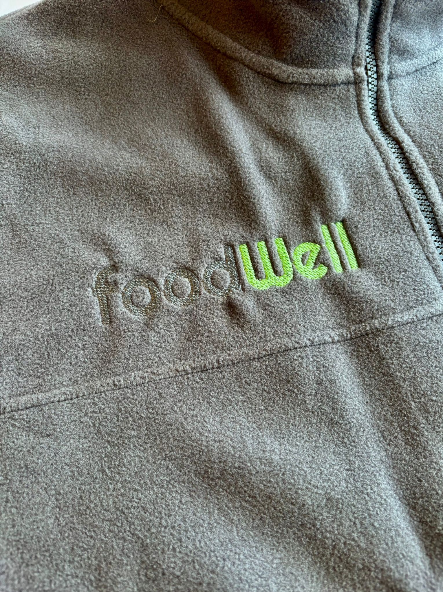 Szary polar z suwakiem i haftowanym, zielonym logo 'foodWell'. Detal haftu, widoczna faktura materiału i zamek błyskawiczny.