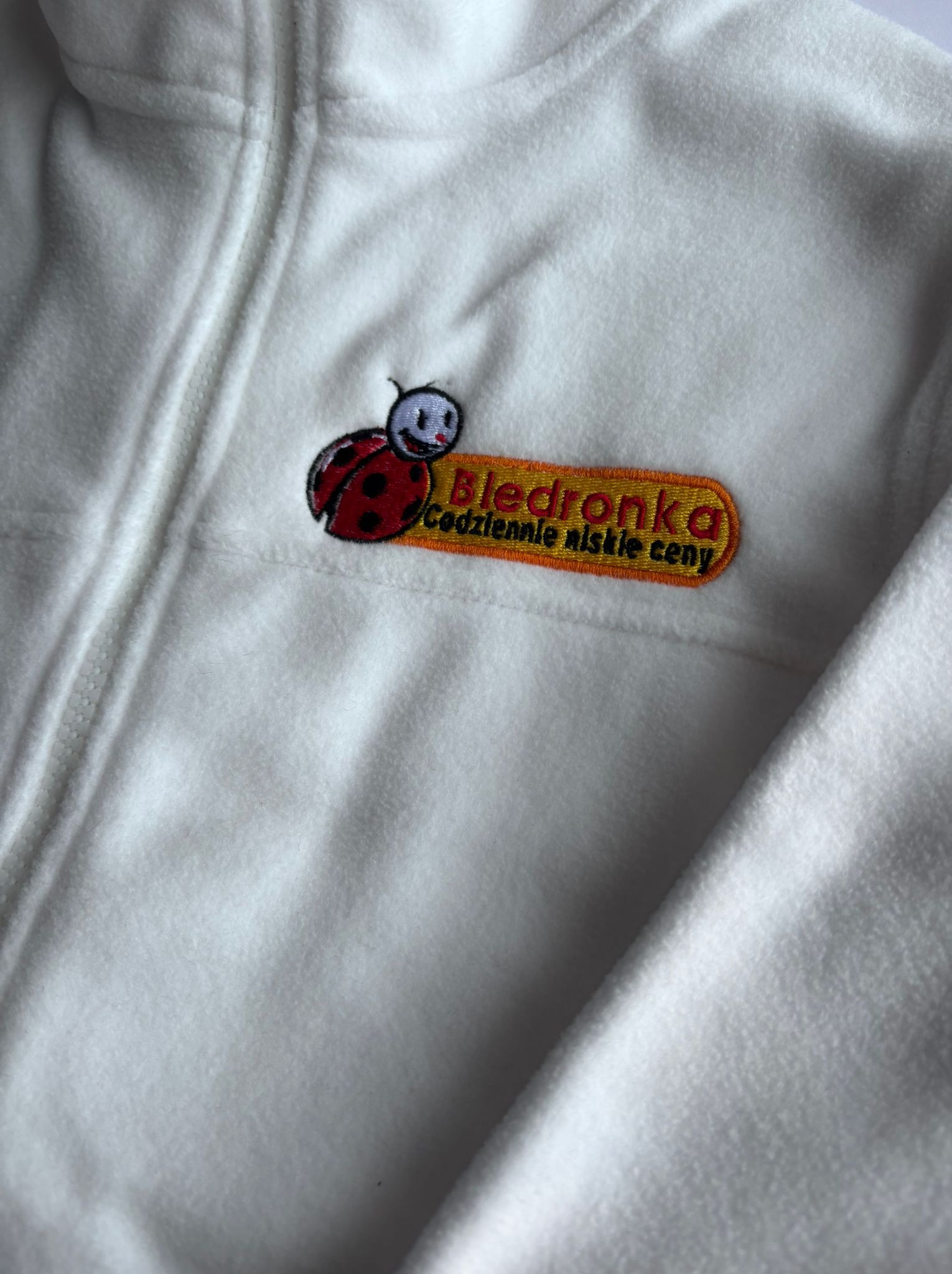 Biała bluza polarowa z haftowanym logo Biedronki (biedronka i napis 'Codziennie niskie ceny') na piersi, widoczne zapięcie na zamek błyskawiczny.
