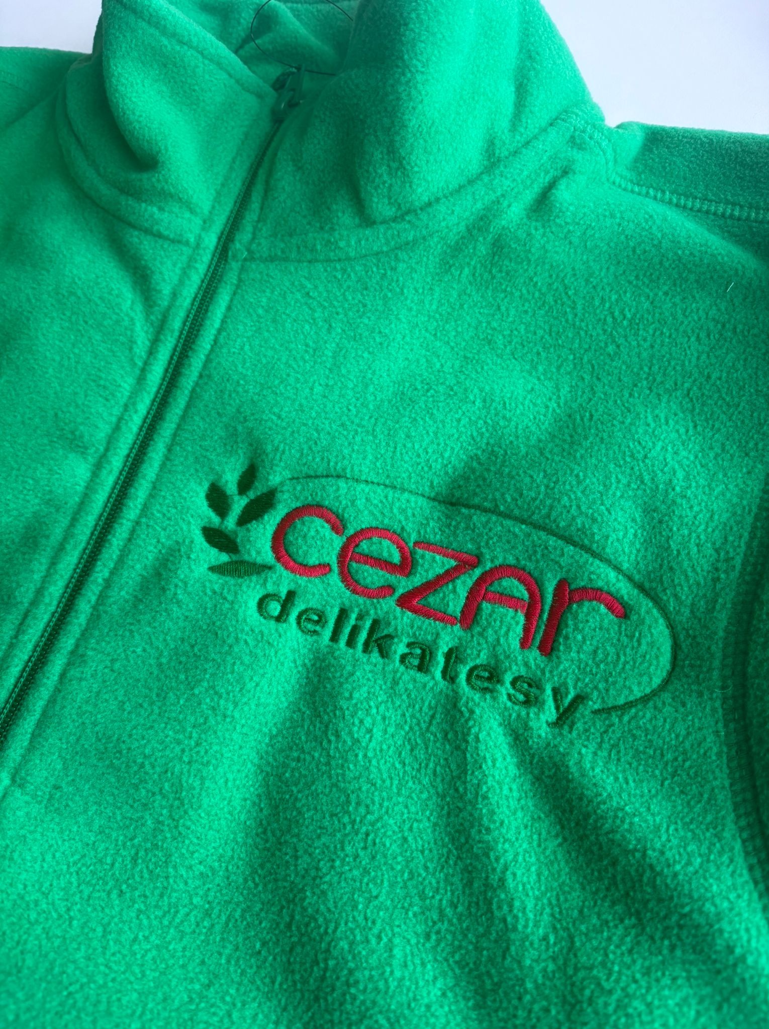 Zielony polar z haftowanym logo 'Cezar delikatesy' w czerwonej obwódce, z motywem roślinnym po lewej stronie. Widoczny zamek błyskawiczny.