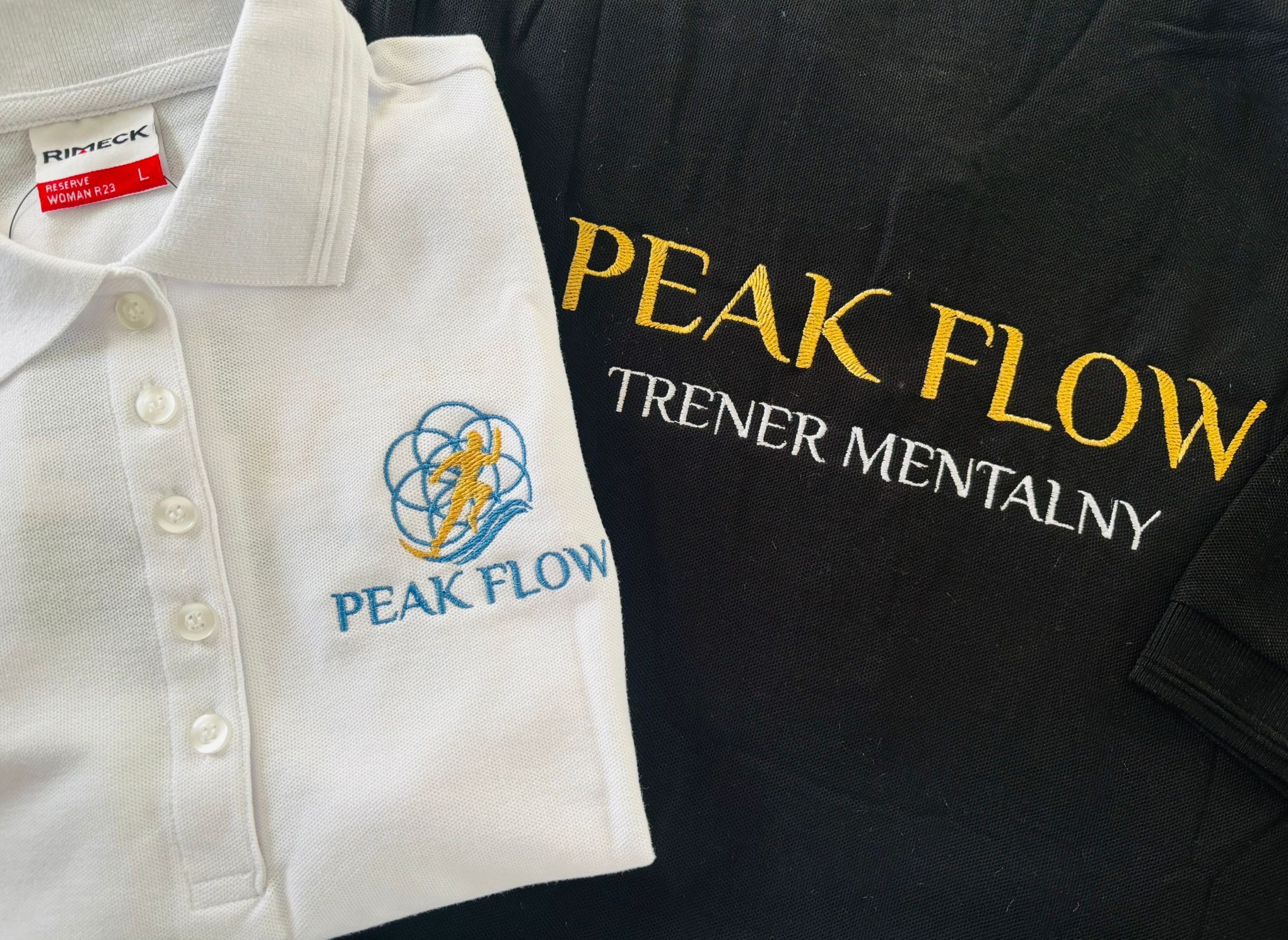 Dwie koszulki polo: biała z niebiesko-złotym logo i czarna z żółto-białym napisem 'PEAK FLOW TRENER MENTALNY'. Widoczny fragment metki Rimeck.