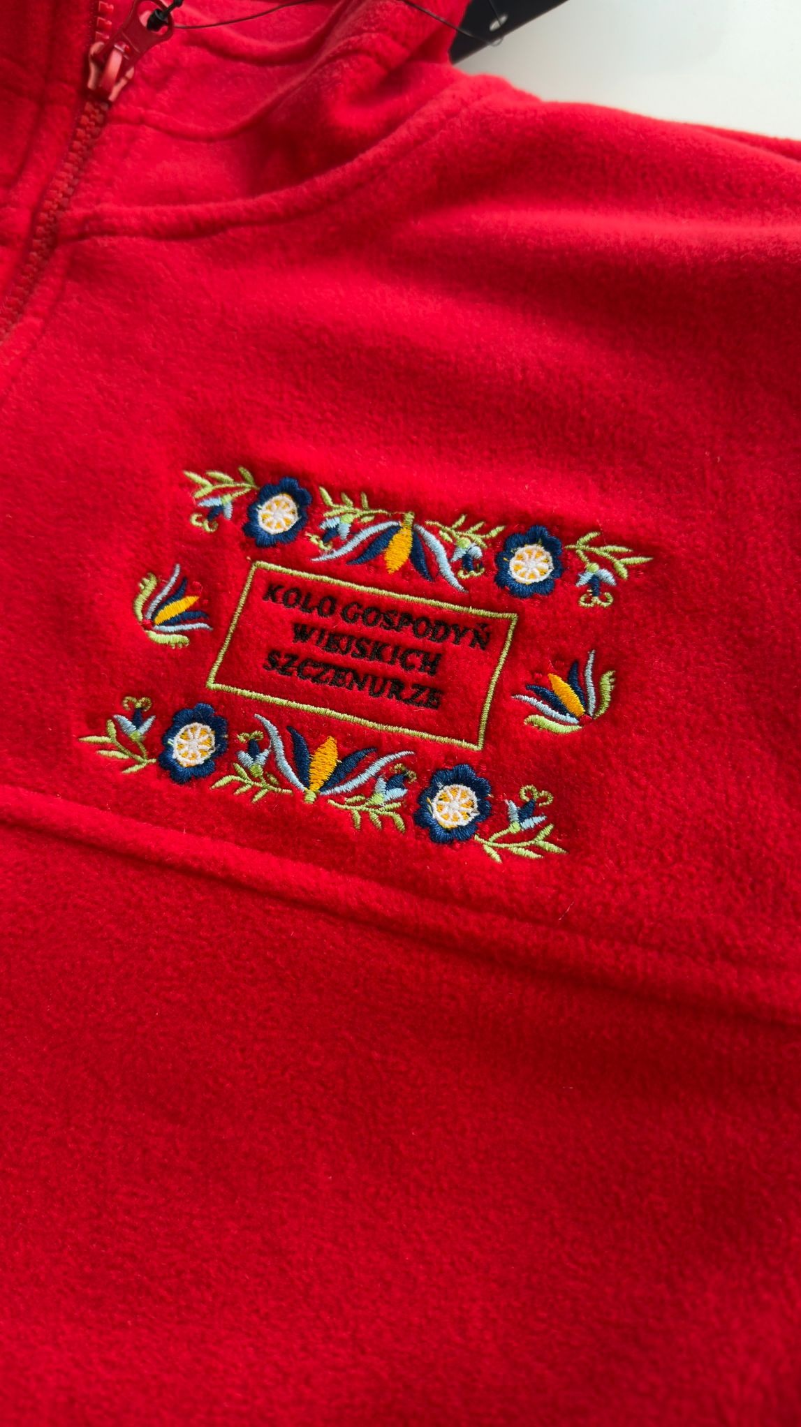 Czerwony polar z haftowanym logo 'Koło Gospodyń Wiejskich Szczawnica', zdobiony motywami kwiatowymi i roślinnymi. Widoczny zamek błyskawiczny.