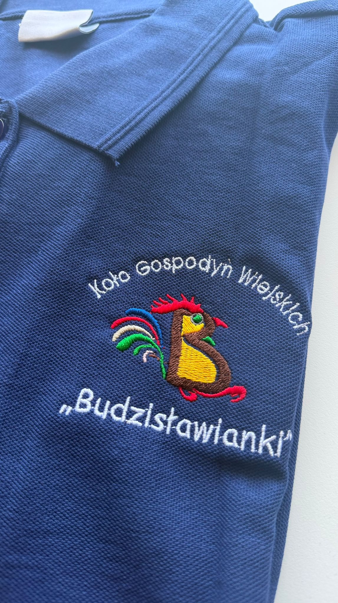Granatowa koszulka polo z kolorowym haftem: kogut, napis 'Koło Gospodyń Wiejskich' i nazwa 'Budzisławianki'. Widoczny kołnierzyk i guziki.