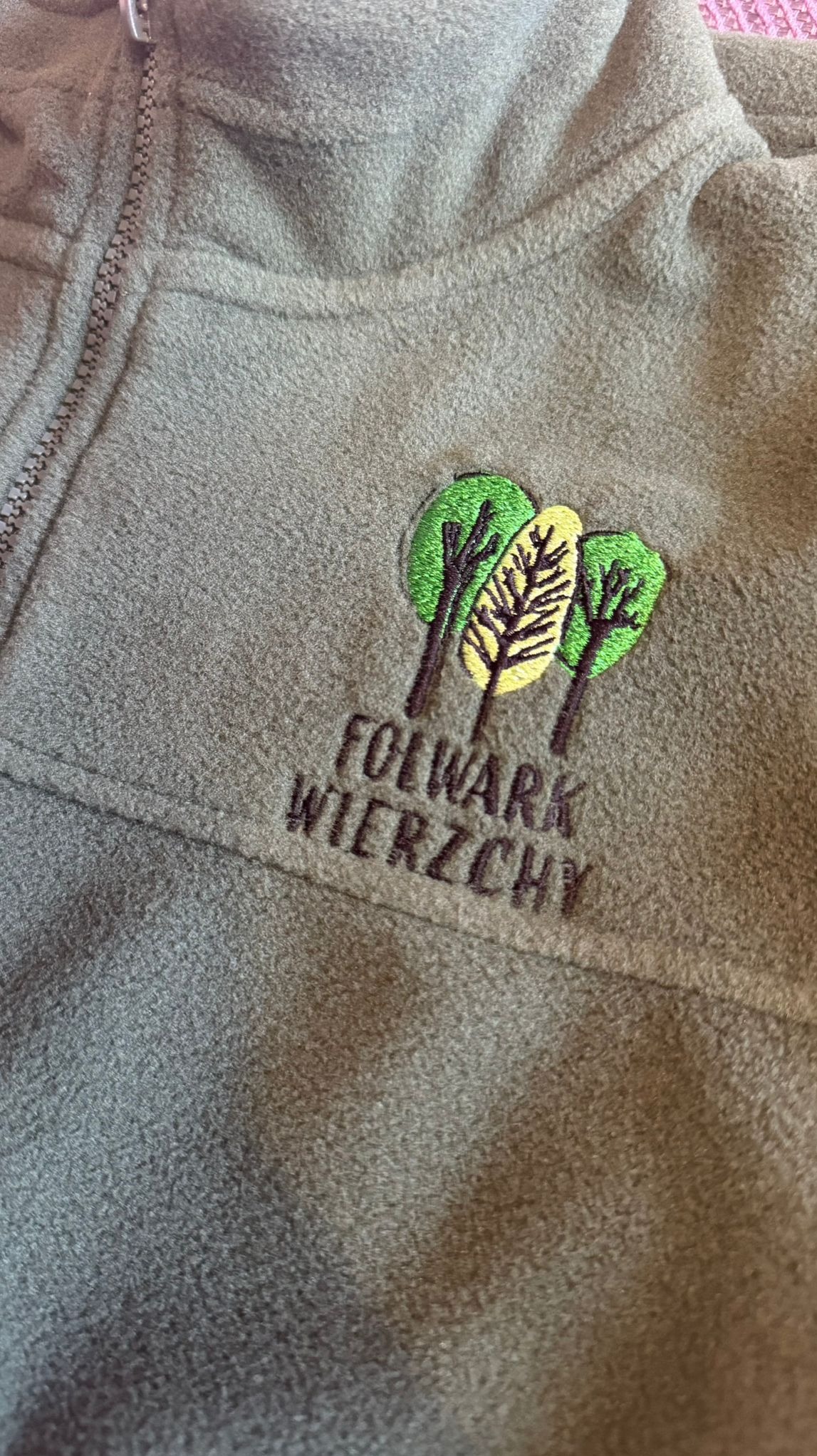 Oliwkowy polar z haftowanym logo firmy 'Folwark Wierzchy' przedstawiającym trzy drzewa, z czego środkowe jest żółte. Widoczny zamek błyskawiczny.