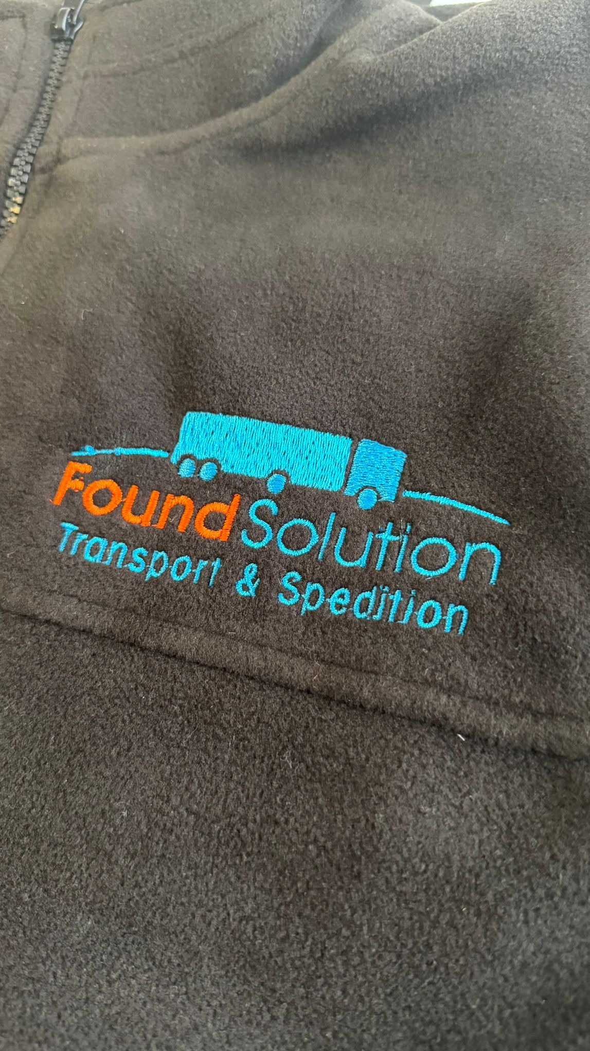 Czarny polar z wyhaftowanym, kolorowym logo firmy transportowej: pomarańczowy napis 'Found Solution' i turkusowy rysunek ciężarówki z naczepą.