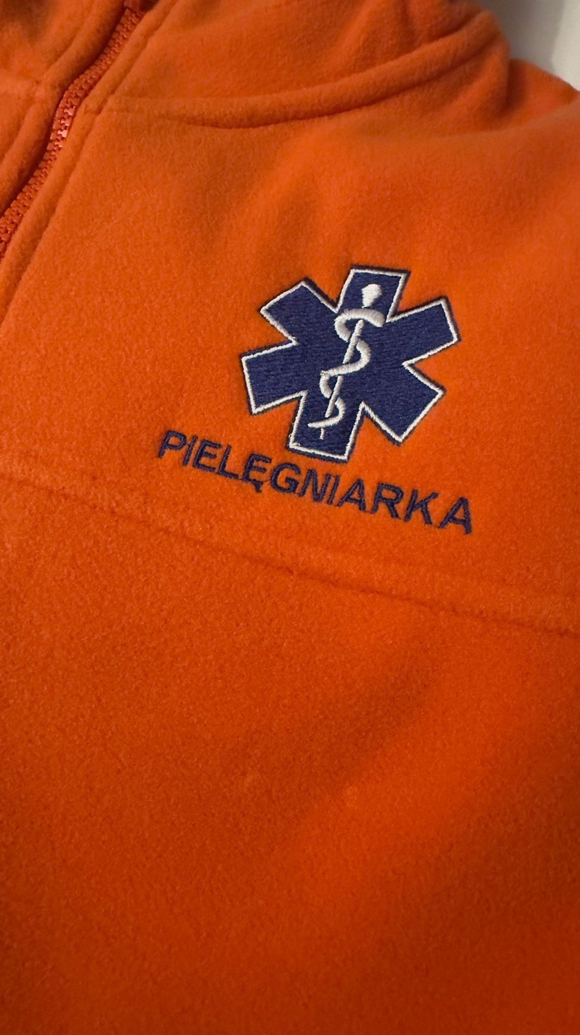 Pomarańczowy polar z wyhaftowanym symbolem medycznym i napisem 'PIELĘGNIARKA' w kolorze granatowym. Widoczne detale zamka błyskawicznego.