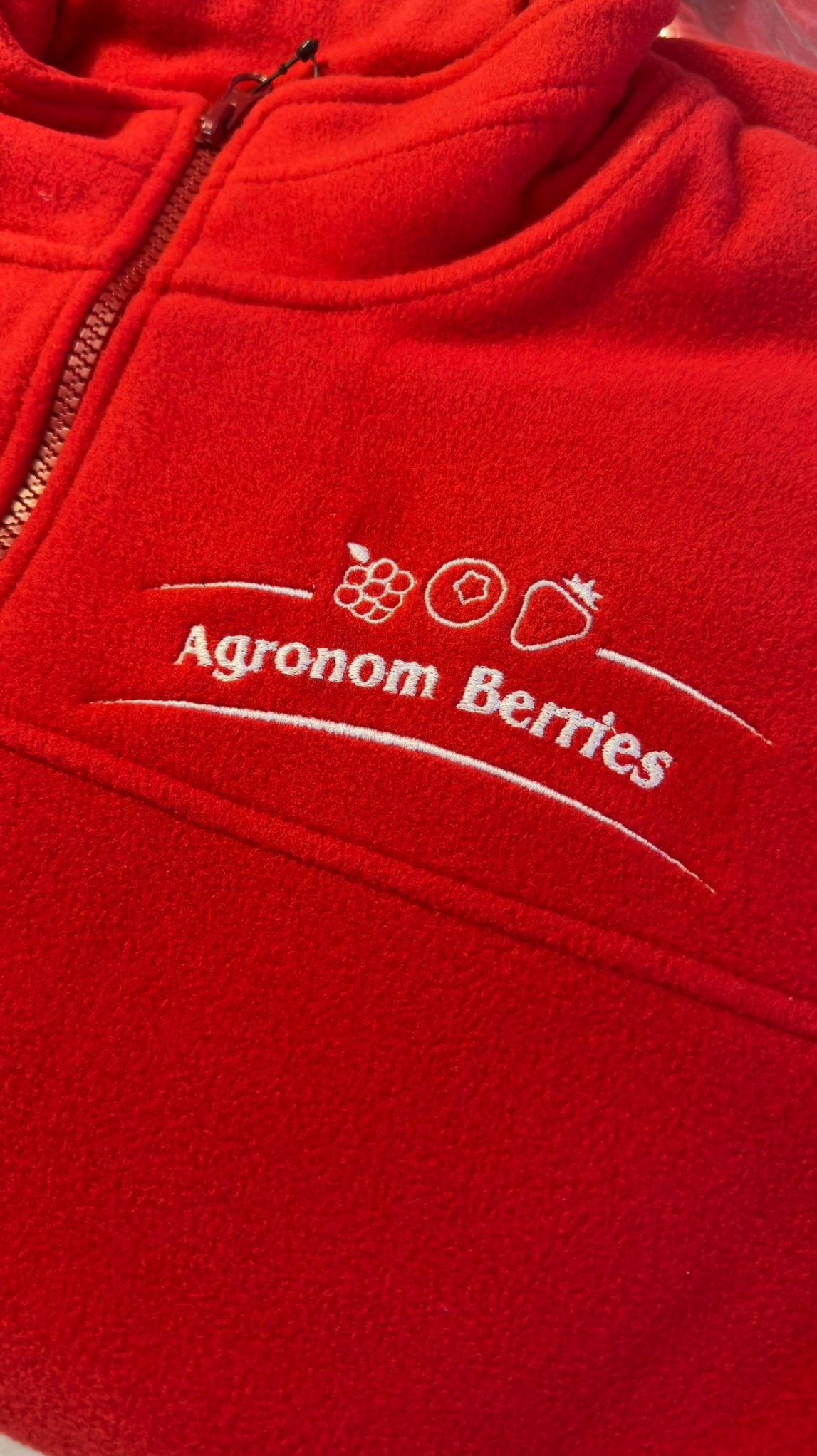 Czerwony polar z białym, precyzyjnym napisem 'Agronom Berries' i motywami owoców. Widoczny zamek błyskawiczny i faktura materiału.