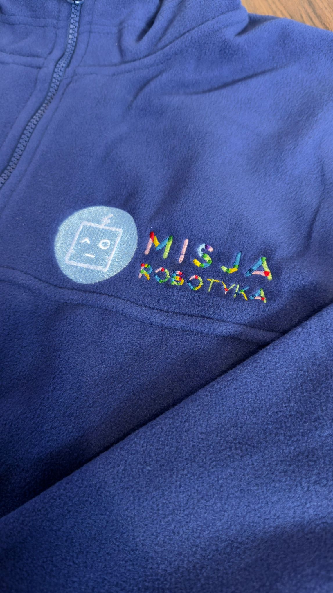 Granatowy polar z suwakiem, ozdobiony kolorowym haftem 'Misja Robotyka' i logo z robotem. Widoczny fragment materiału i zamek błyskawiczny.