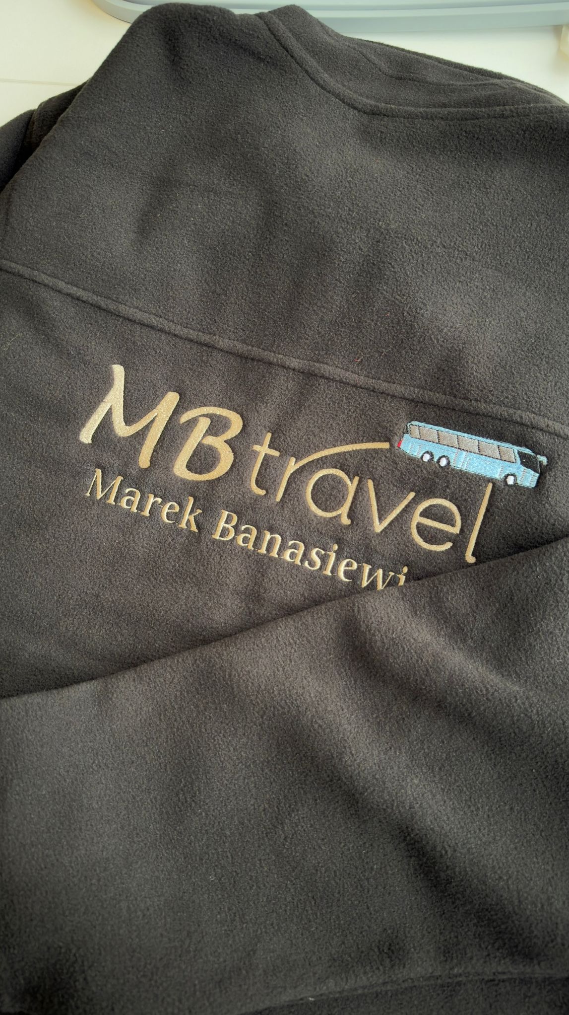 Grafitowy polar z wyhaftowanym złotym logo 'MBtravel' i niebieskim autobusem, imię i nazwisko pod spodem. Widoczne delikatne zmarszczenia materiału.