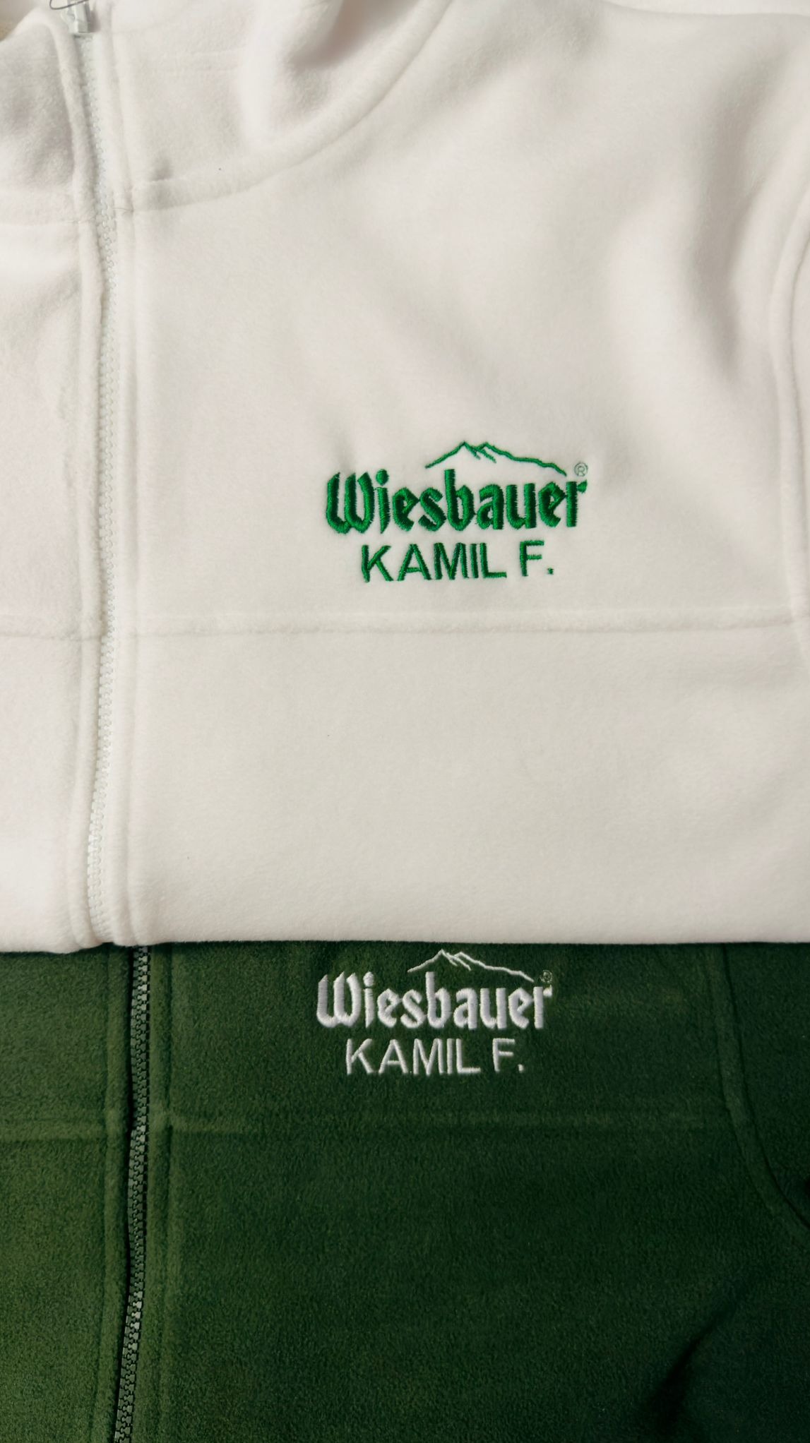 Dwa polarowe swetry z haftowanym logo firmy Wiesbauer i imieniem Kamil F., jeden biały z zielonym haftem, drugi zielony z białym haftem. Widoczne zamki błyskawiczne.