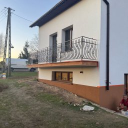 Fliz-kam - Dwukondygnacyjny dom z balkonem z ozdobną, metalową balustradą, dolna część elewacji w kolorze brązowym, górna biała, widoczny pracownik w trakcie wykonywania prac wykończeniowych przy elewacji.