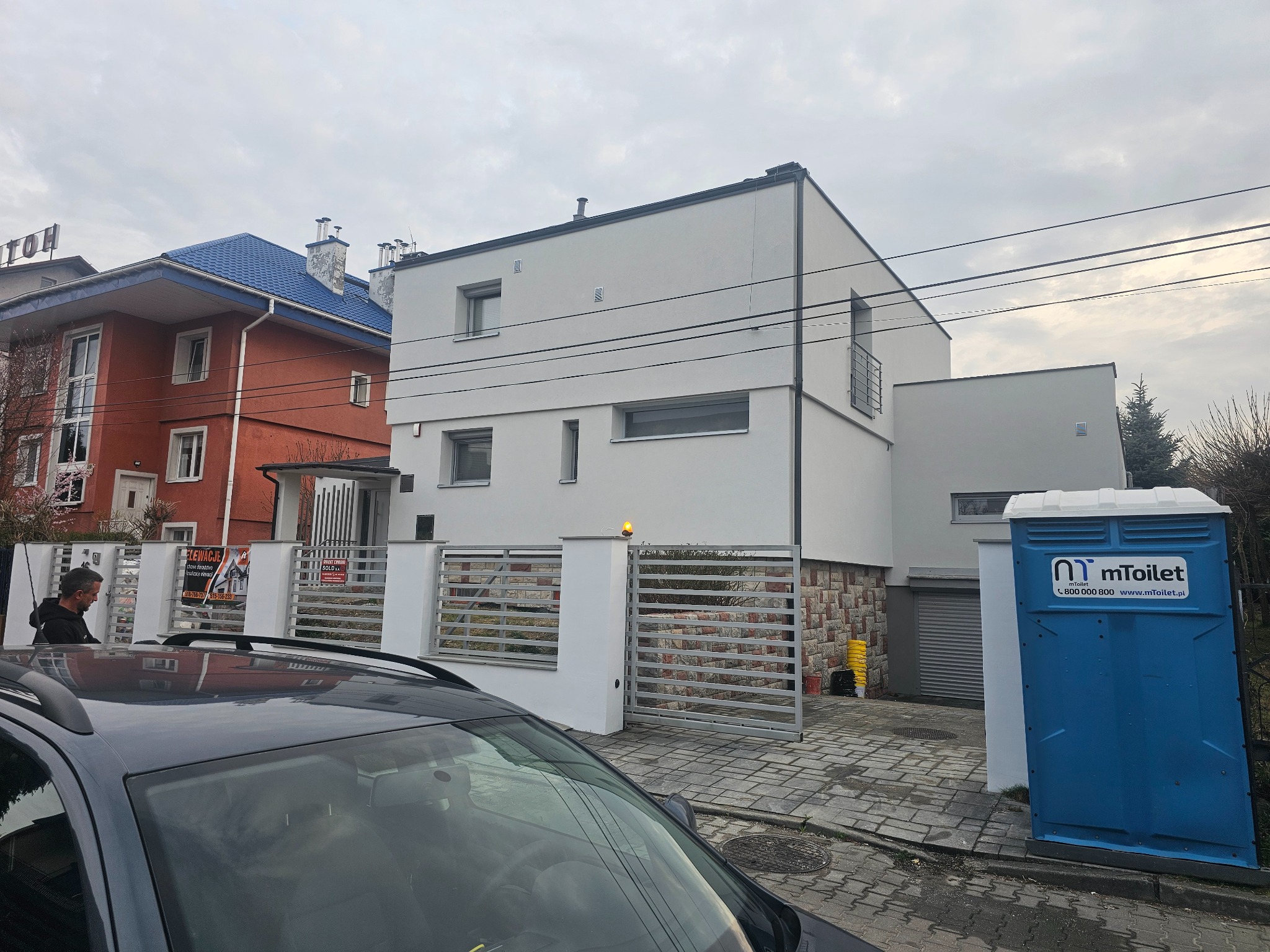 Nowoczesny, biały dom jednorodzinny z elementami kamiennej elewacji na cokole, widoczna brama wjazdowa i przenośna toaleta na posesji, pochmurne niebo w tle.