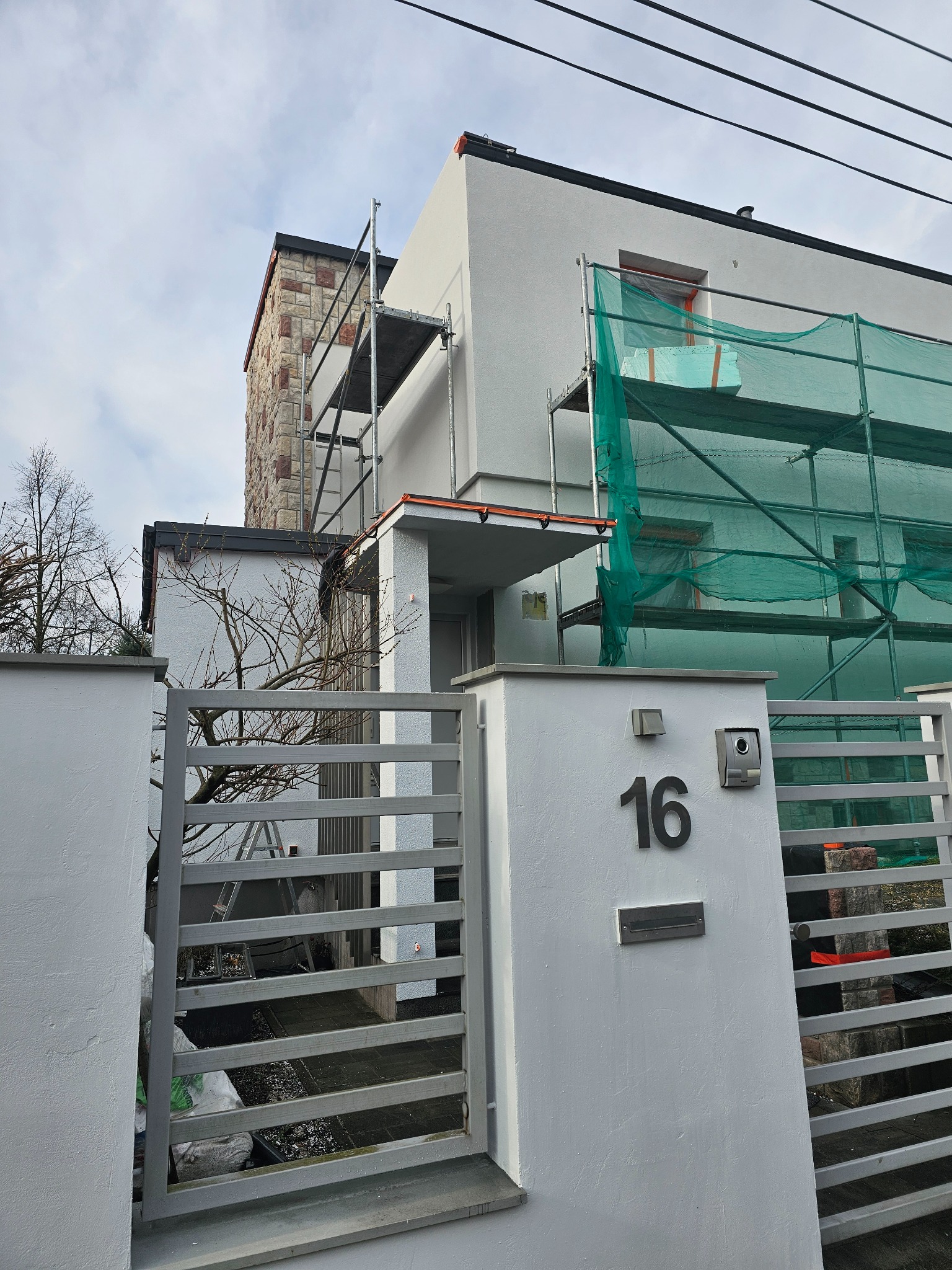 Nowoczesny dom z białymi ścianami i kamienną fasadą w trakcie prac elewacyjnych, widoczne rusztowania i siatka ochronna, numer domu '16' na ogrodzeniu.