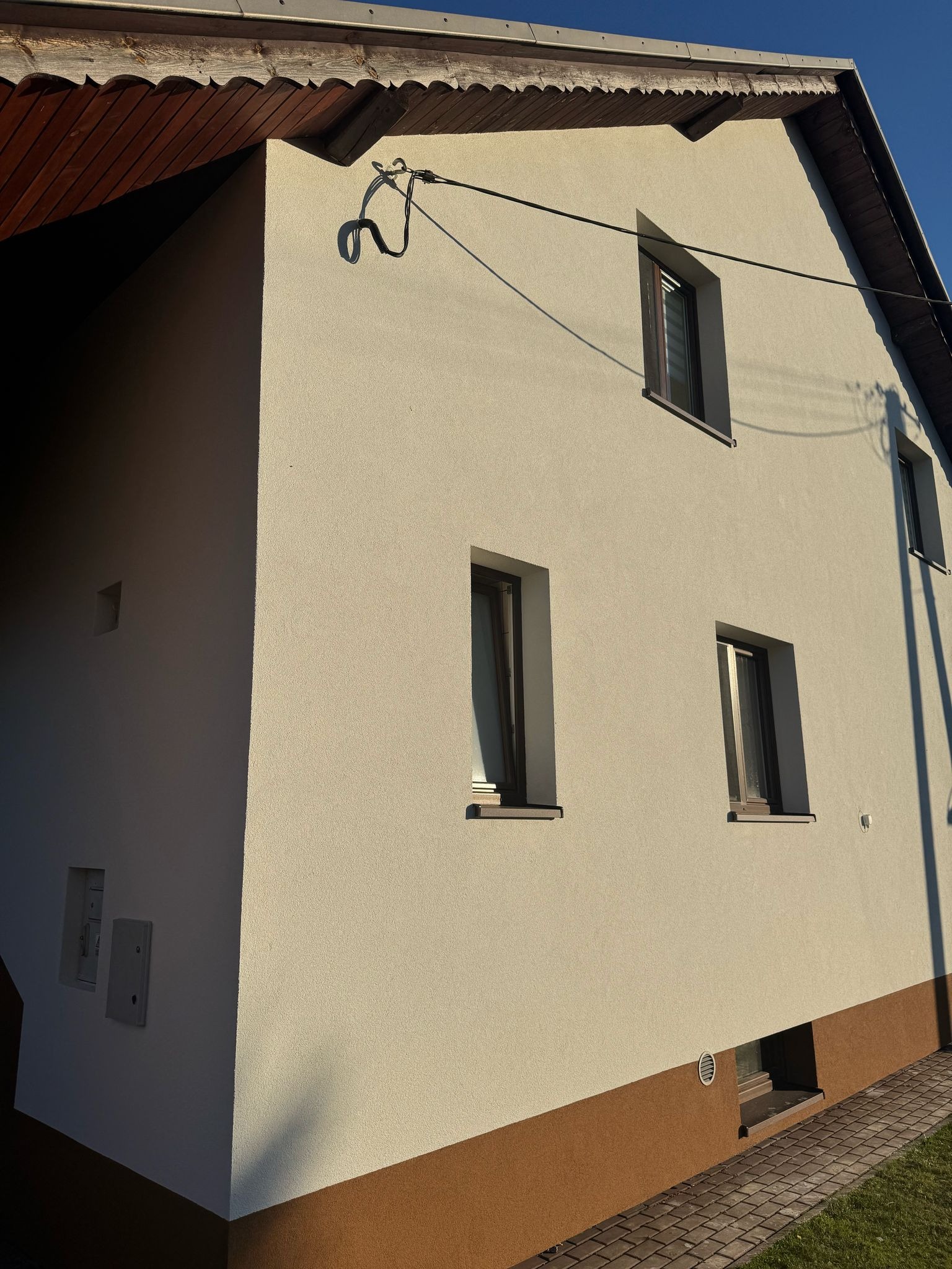Jasna elewacja domu jednorodzinnego z brązowym pasem u dołu, widoczne trzy okna z brązowymi ramami, kabel elektryczny i fragment dachu z drewnianą podbitką.