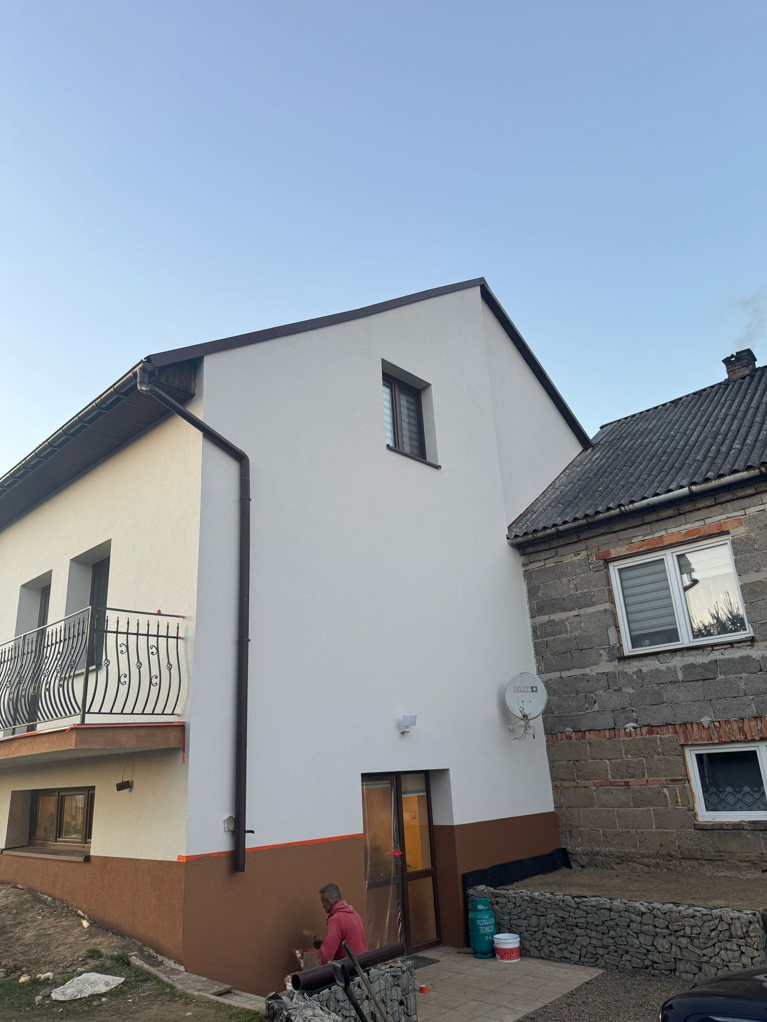 Dwukondygnacyjny dom po remoncie elewacji, z białym tynkiem na górze i brązowym na dole, obok starszy budynek z szarej cegły, widoczny pracownik malujący dolną część elewacji na brązowo.