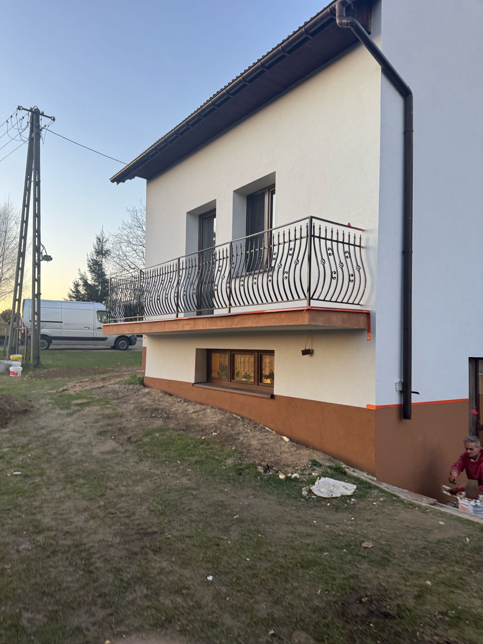 Dwukondygnacyjny dom z balkonem z ozdobną, metalową balustradą, dolna część elewacji w kolorze brązowym, górna biała, widoczny pracownik w trakcie wykonywania prac wykończeniowych przy elewacji.