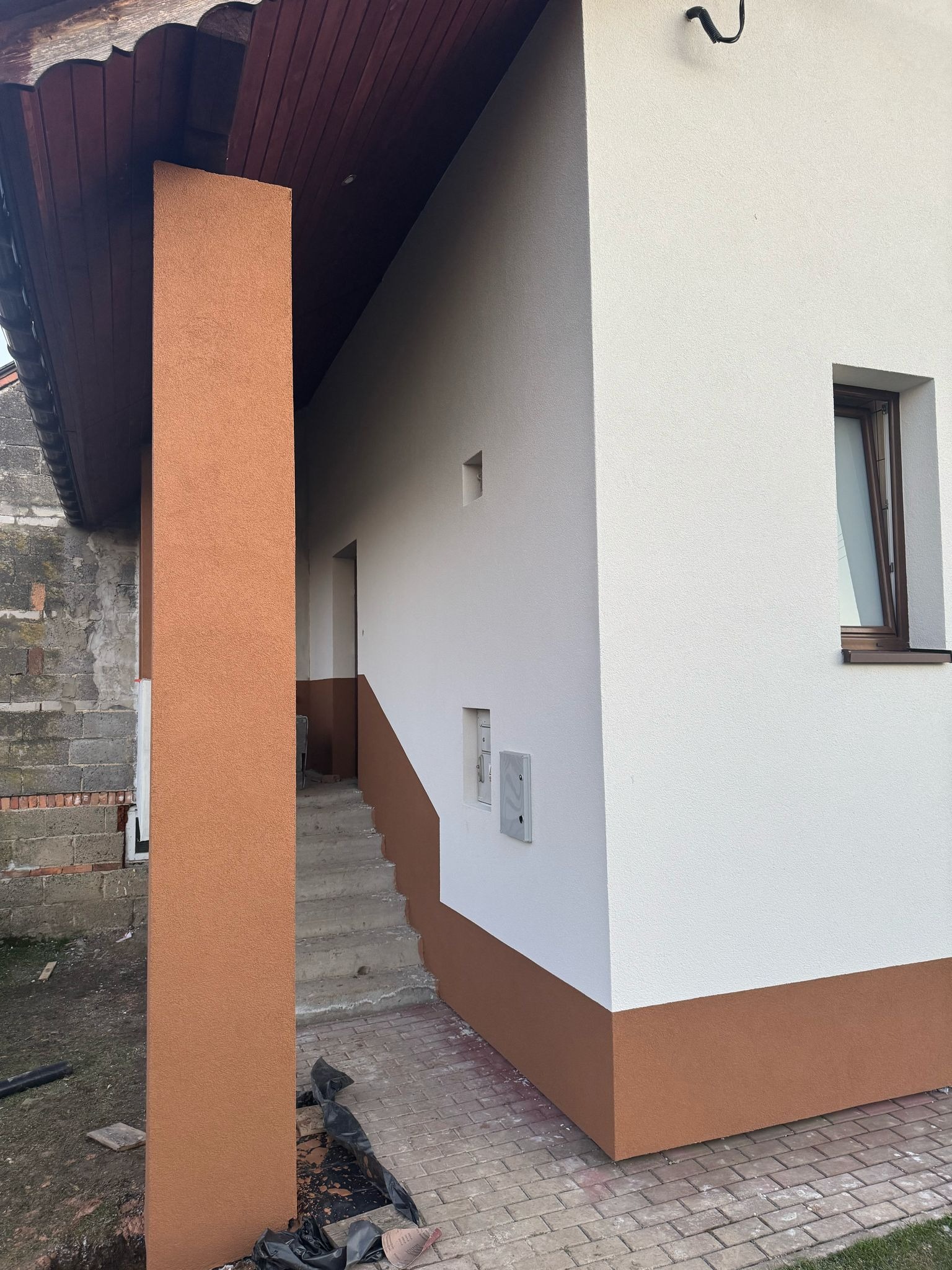 Świeżo otynkowana elewacja domu z widocznym fragmentem schodów i brązowym cokołem, kontrastującym z białą ścianą. Widoczny słupek w kolorze cokołu.
