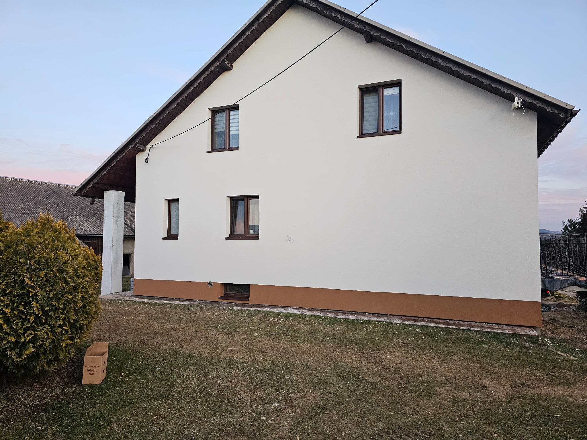 Dwukondygnacyjny dom z nową, białą elewacją i brązowym cokołem, widoczny z perspektywy trawnika z kartonem po prawej stronie, na tle nieba o zmierzchu.