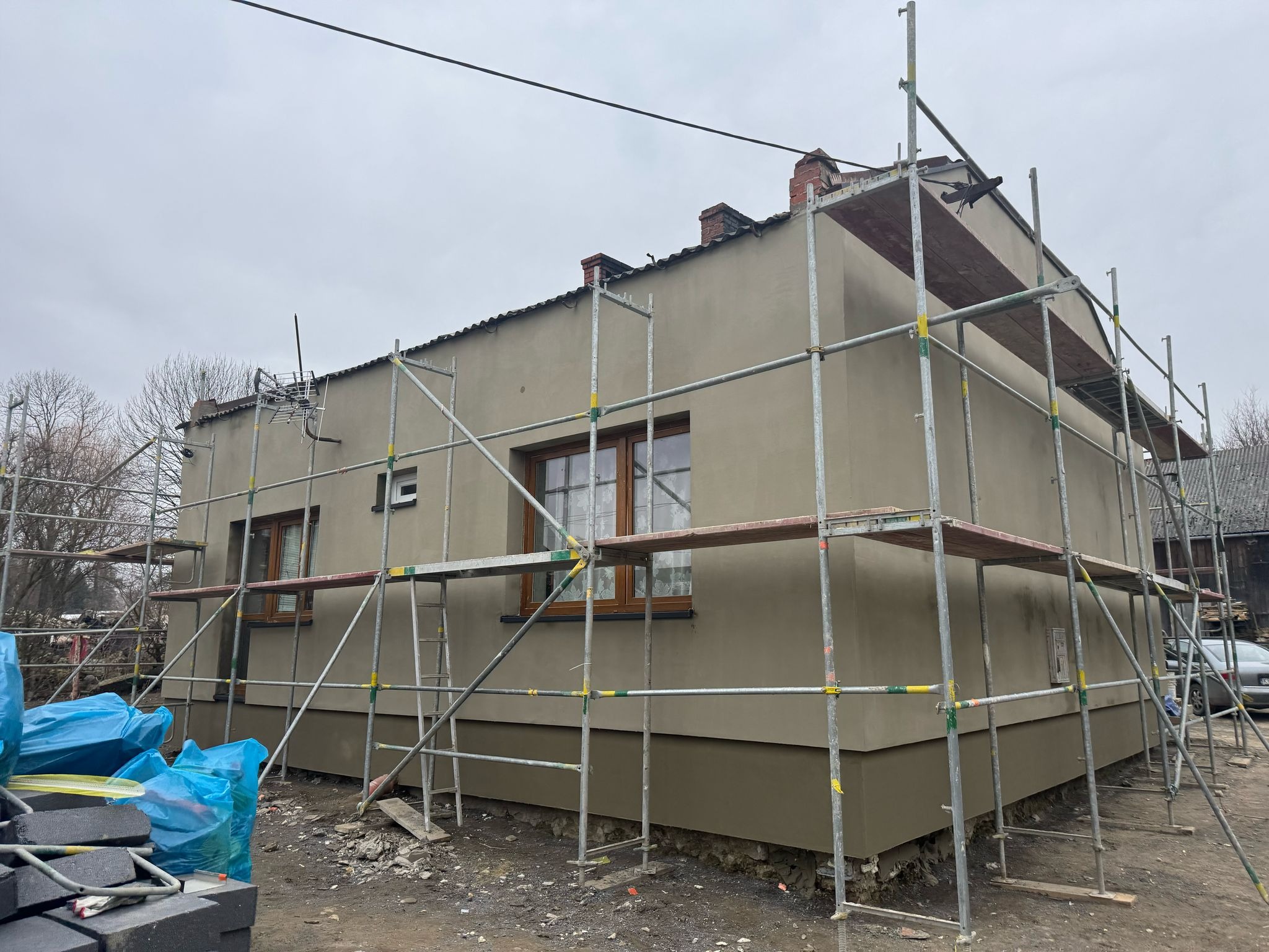 Dom jednorodzinny ze świeżo nałożonym tynkiem elewacyjnym w odcieniach szarości, otoczony rusztowaniem; widoczne okna z drewnianą stolarką, stos materiałów budowlanych w niebieskich workach...
