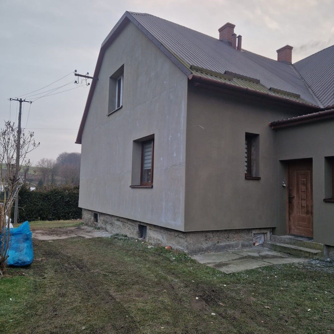 Dom jednorodzinny z szarą, surową elewacją, widoczne okna z roletami, brązowe rynny i metalowy dach, teren wokół domu częściowo porośnięty trawą, pochmurne niebo.