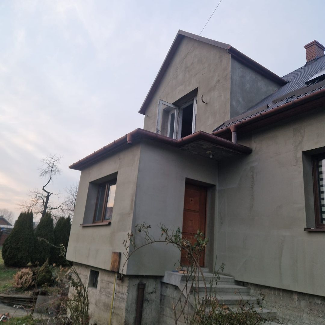 Dom jednorodzinny z szarą elewacją w trakcie prac, widoczne świeżo otynkowane ściany, otwarte okno na poddaszu, brązowe rynny i schody wejściowe.