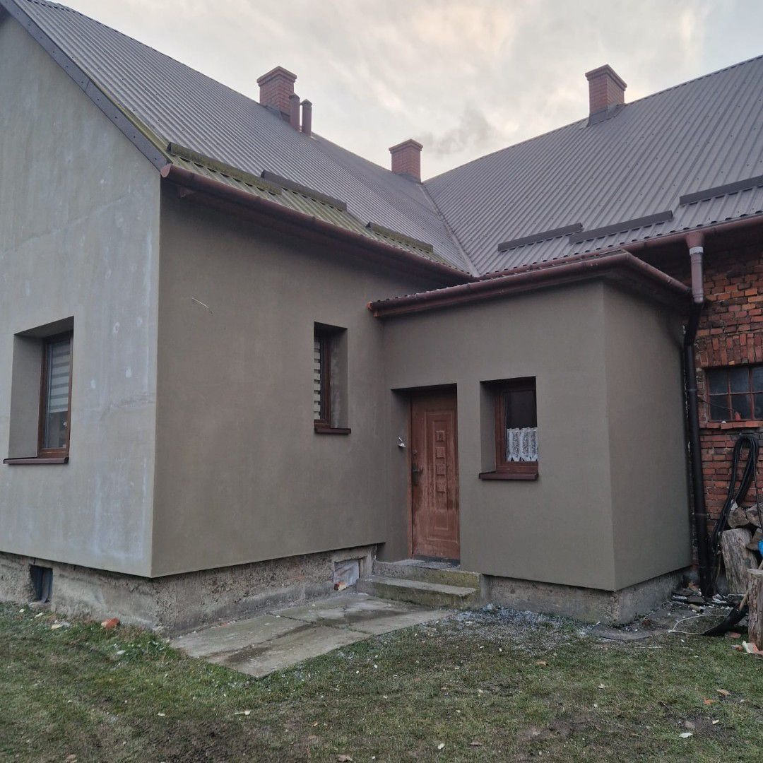 Dom jednorodzinny z szarą, świeżo nałożoną elewacją, widoczna stara ceglana ściana z boku, brązowe rynny i metalowy dach, pochmurne niebo w tle.