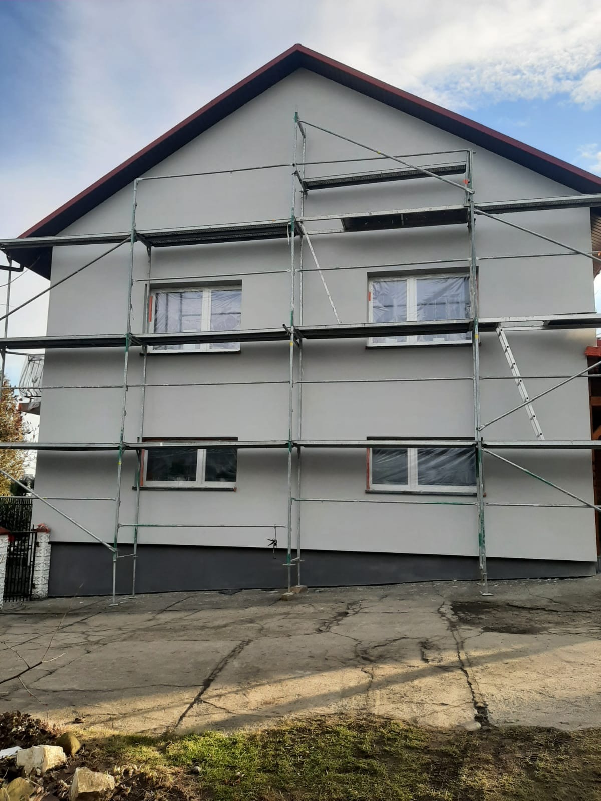 Dwukondygnacyjny dom jednorodzinny z szarą elewacją, na której zamontowane są rusztowania, okna zabezpieczone folią, cokół pomalowany na ciemny grafit.