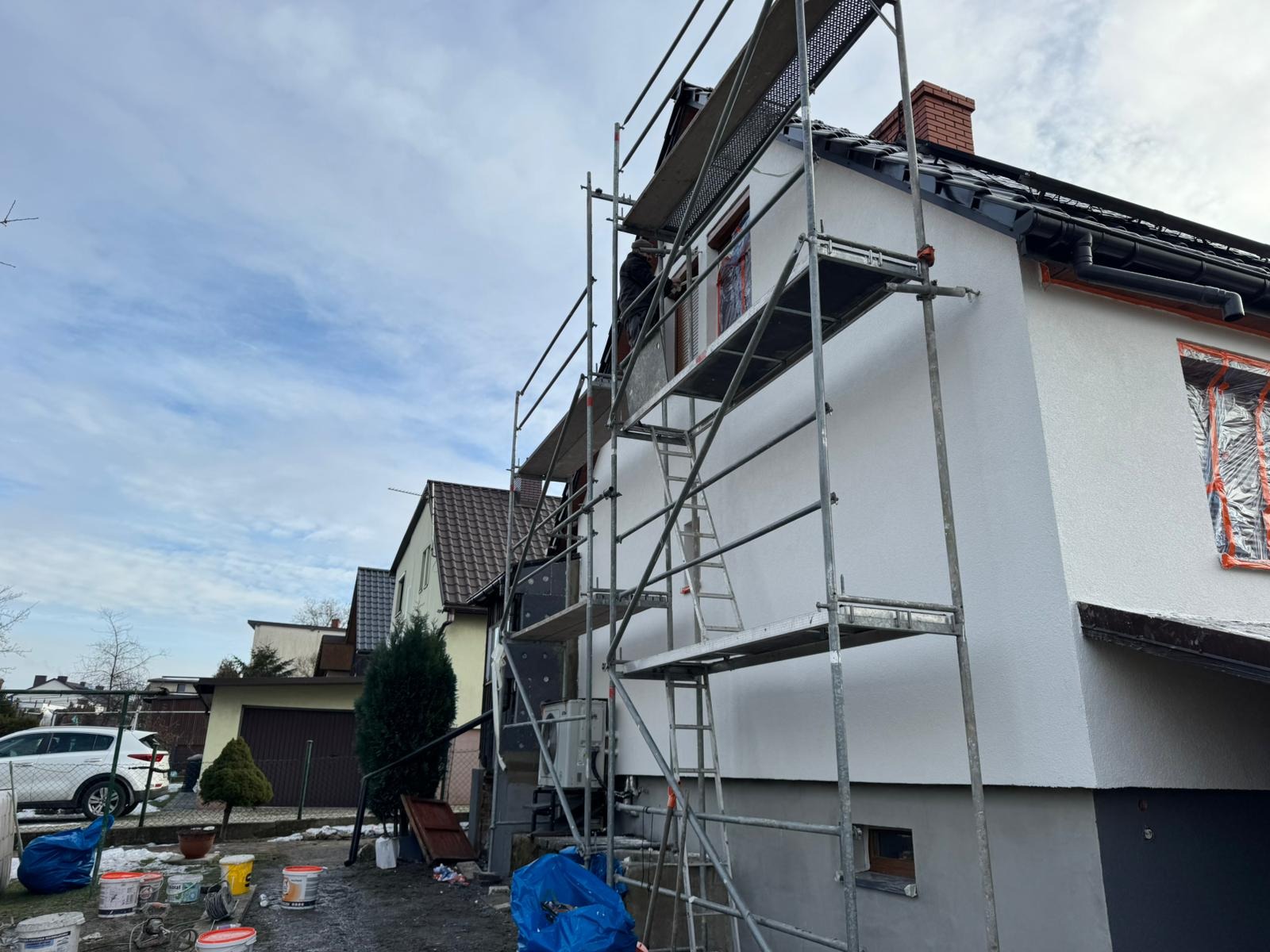 Ocieplony dom w trakcie tynkowania elewacji, widoczne rusztowania, zabezpieczone okna folią, częściowo ocieplona ściana z szarym tynkiem strukturalnym, przygotowane farby i narzędzia malarskie.