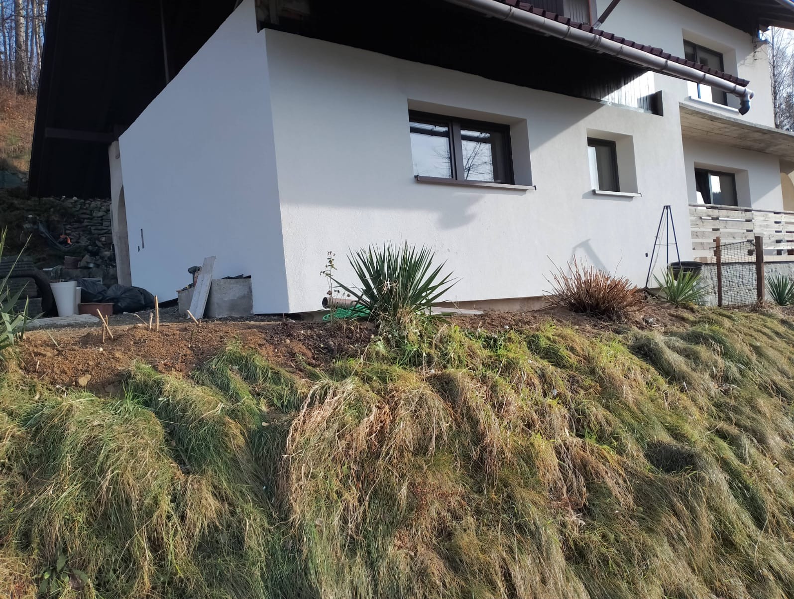 Biała elewacja domu jednorodzinnego z brązowymi ramami okiennymi, widoczna z perspektywy zbocza porośniętego trawą i roślinnością.