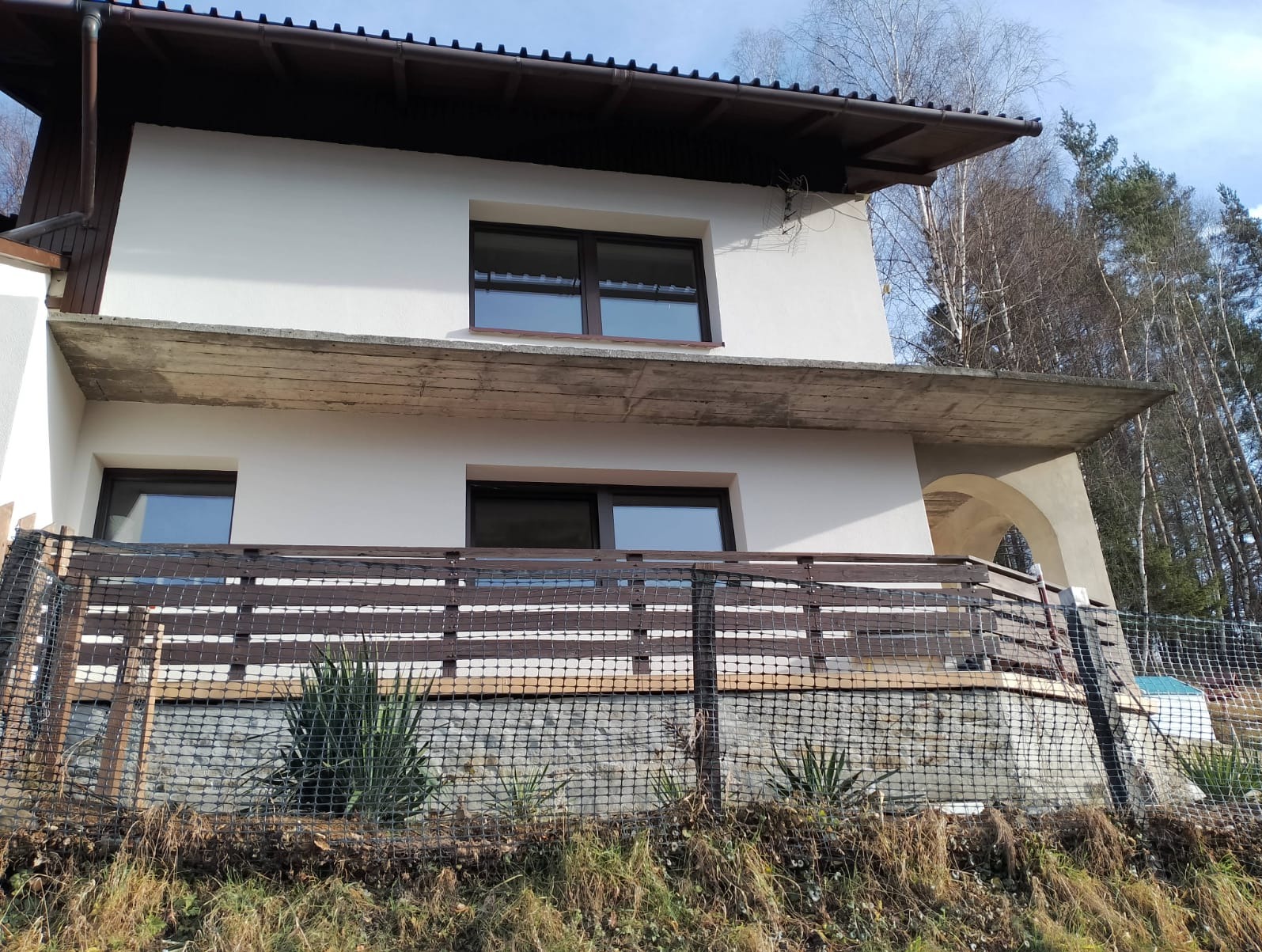 Dwupiętrowy dom z jasną elewacją, brązowymi oknami i drewnianą balustradą balkonu, widoczny przez siatkę ochronną. Dom otoczony drzewami, ujęcie z dołu.