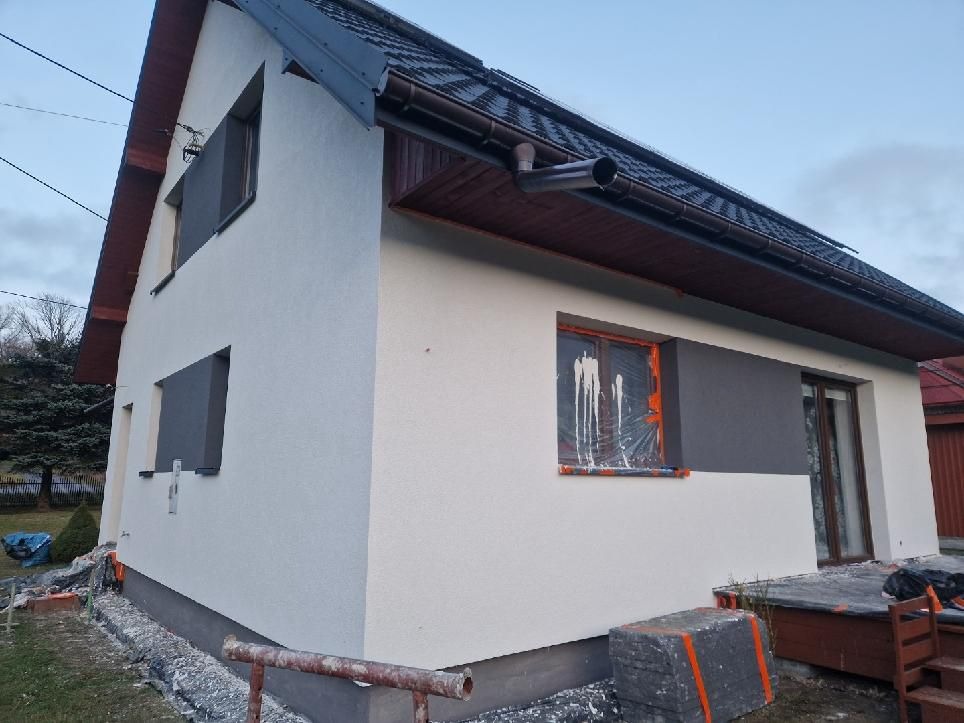 Dwukondygnacyjny dom po tynkowaniu elewacji, z częściowo zabezpieczonym oknem folią malarską i widocznymi pozostałościami po pracach wykończeniowych wokół budynku.