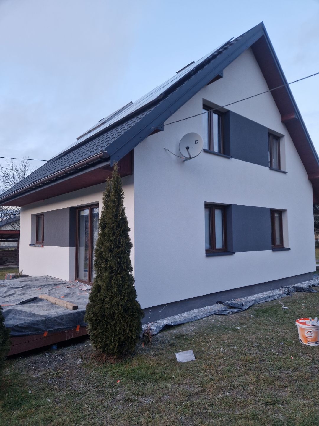 Dwukondygnacyjny dom jednorodzinny z nową, dwukolorową elewacją w odcieniach bieli i szarości, widok z boku, z widoczną anteną satelitarną i panelami słonecznymi na dachu.