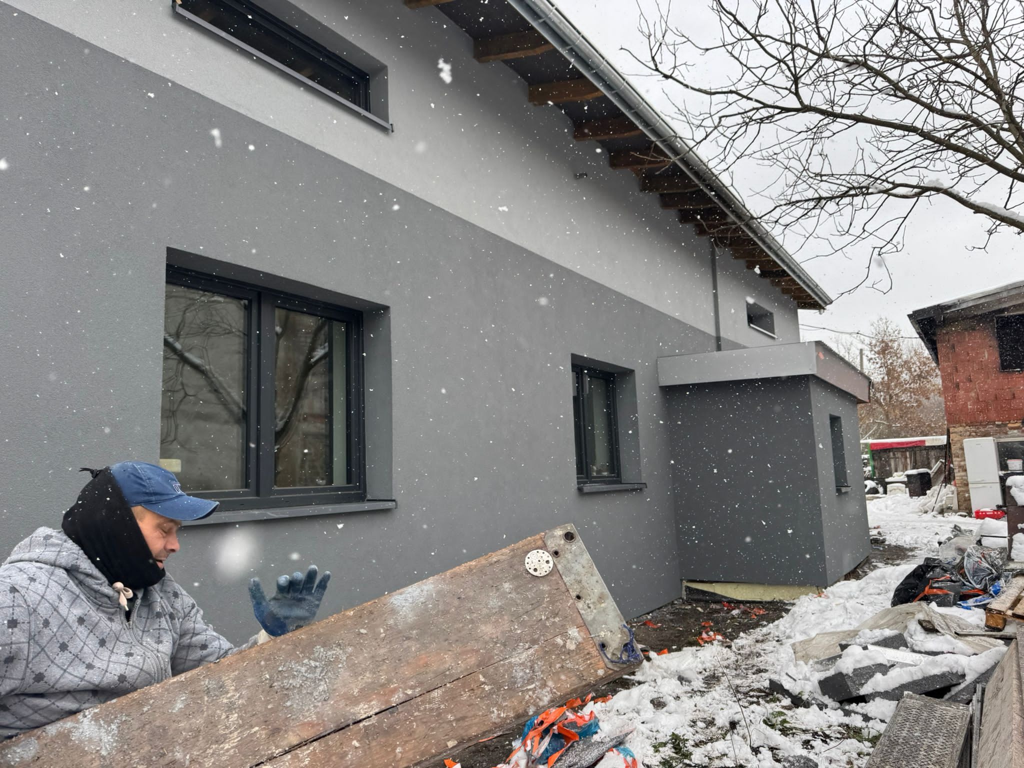 Pracownik w roboczym ubraniu trzyma deskę na tle otynkowanej szarej elewacji domu podczas opadów śniegu.