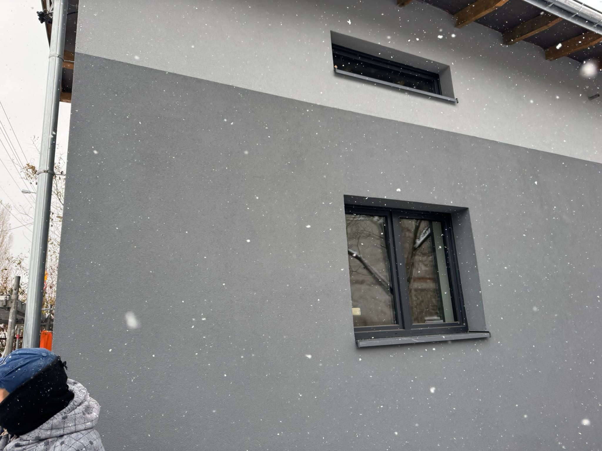 Fragment dwukolorowej elewacji budynku z dwoma oknami, sfotografowany podczas opadów śniegu.