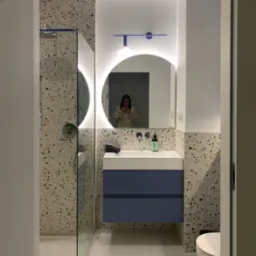 Nowoczesna łazienka z kabiną prysznicową za szkłem, umywalką z szafką w kolorze granatowym, okrągłym lustrem z podświetleniem LED i płytkami imitującymi lastryko na ścianach; w lustrze odbija się...