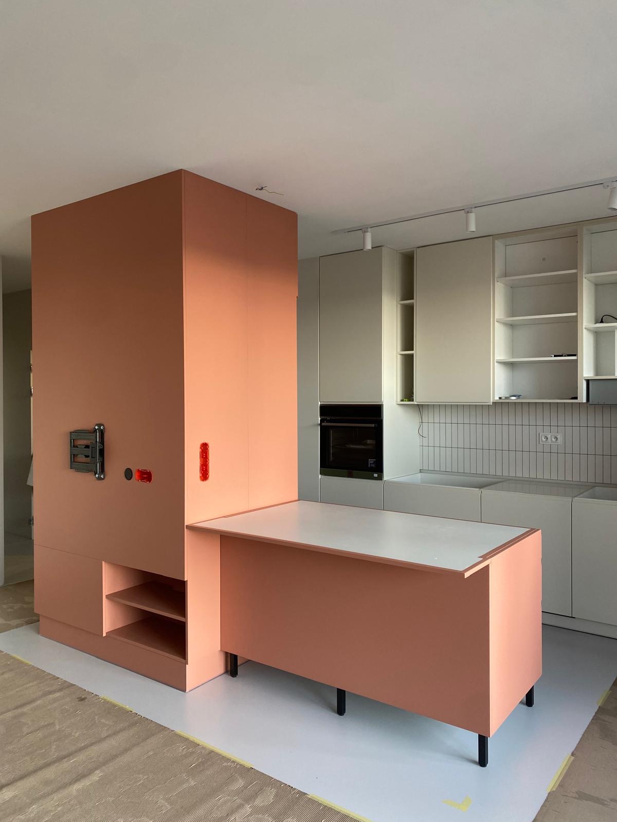 Wnętrze kuchni w trakcie montażu: wysoka zabudowa w odcieniach brzoskwini z elementami instalacji elektrycznej, biały blat roboczy i szare szafki kuchenne z piekarnikiem w tle, widoczne...