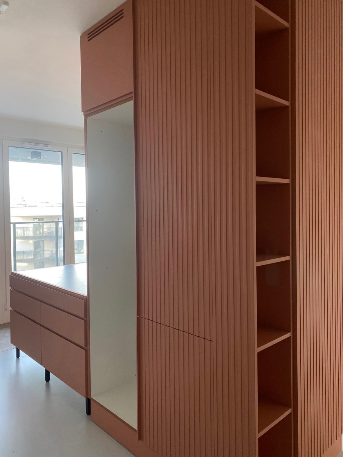 Mieszkanie w Poznaniu: zabudowa meblowa w odcieniach terakoty z ryflowanymi frontami szaf i szuflad, widoczne półki i lustro, w tle okno z widokiem na balkon.