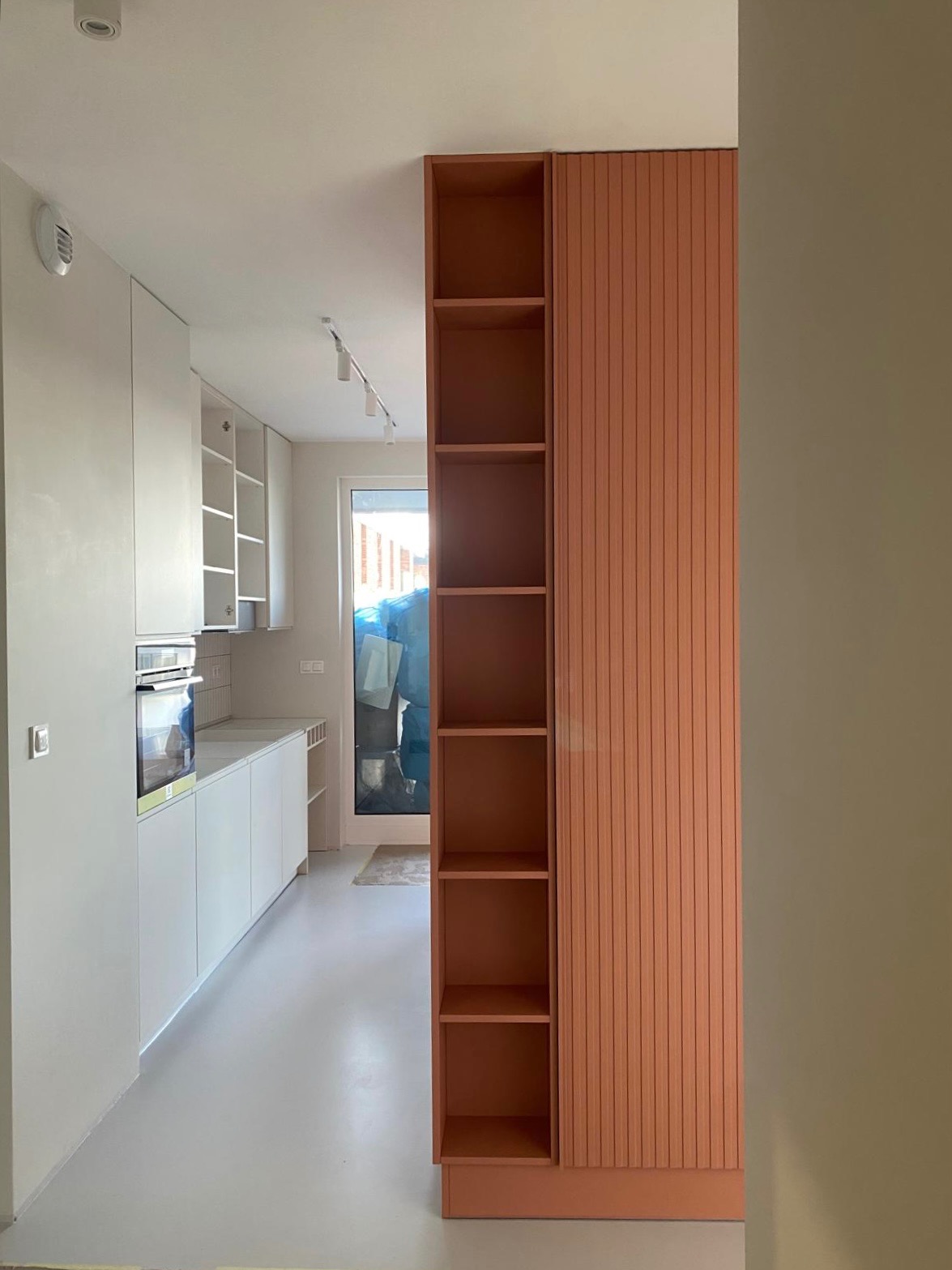 Wnętrze kuchni w trakcie wykańczania: białe szafki kuchenne, piekarnik w zabudowie, płytki na ścianie nad blatem, terakotowy regał z półkami i lamelami, widok na drzwi balkonowe z folią ochronną.