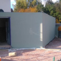 MUR-BETON - Nowo otynkowany budynek gospodarczy w trakcie wykańczania. Widoczne zabezpieczenia folią, drabina i materiały budowlane. Prace wykończeniowe na zewnątrz.