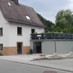 Dwukondygnacyjny dom z balkonem i kamienną elewacją do połowy wysokości. Widoczne szare tynki, brązowy dach i kostka brukowa na podjeździe.