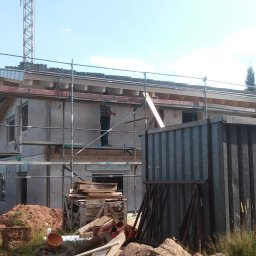 MUR-BETON - Budowa domu jednorodzinnego w trakcie realizacji: widoczne rusztowania, ściany z bloczków, częściowo zadaszony dach. Na rusztowaniu stoi osoba w kraciastej koszuli.