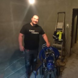 MUR-BETON - Uśmiechnięty mężczyzna w roboczym ubraniu stoi obok profesjonalnego agregatu malarskiego w surowym wnętrzu, przygotowanym do malowania. W tle drabina.