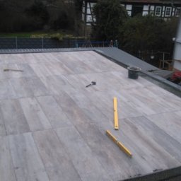 MUR-BETON - Układanie jasnoszarych płytek na dachu tarasu. Widoczne narzędzia: poziomica, młotek gumowy i wiadro. W tle dom z białymi ścianami i zielonymi drzewami.