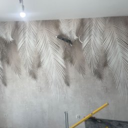 MUR-BETON - Ściana z tapetą w motywie palmowych liści, na dole widoczna poziomica, sugerująca prace wykończeniowe. Oświetlenie sufitowe podkreśla wzór tapety.